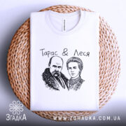 Футболка Тарас і Леся, біла, складена на кошику, ракурс зверху, фон світло-сірий, принт з малюнком Тараса Шевченка та Лесі Українки, текст "Тарас & Леся" над малюнком.