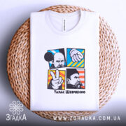 Футболка Тарас Шевченко, біла, на столі, з фоном у світло-сірому кольорі, складена, принт складається з чотирьох квадратів з зображеннями, один з яких містить жест миру, інші - портрети та руки, кольори принта яскраві: синій, червоний, жовтий.