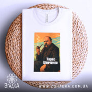 Футболка Тарас Шевченко, біла, на столі, з ракурсу зверху, фон світло-сірий, принт з зображенням Тараса Шевченка з сигаретою, кольори принта: оранжевий, зелений, чорний, текст "Тарас Шевченко" внизу, футболка лежить на плетеній основі.