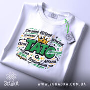 Футболка тато подарунок, біла, на плоскій поверхні, з принтом, що містить слова "ТАТО" та інші позитивні характеристики, фон світлий, деталі принта яскраві, з короною, прикріплена бірка.