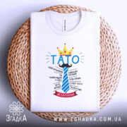 Футболка тато завжди правий, біла, на плоскій поверхні, фон світлий, принт з написом 'ТАТО' у блакитному кольорі, корона жовта, деталі тексту навколо, на плетеній основі.