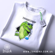 Футболка Травозавр білого кольору, розкладена на поверхні. Принт зображає зеленого динозавра в окулярах, що тримає колбу з блакитною рідиною та пакет з травою. На футболці є етикетка з логотипом, а фон світлий і однорідний.