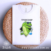 Футболка Травозавр білого кольору, складена на плетеній кошику. Принт зображає зеленого динозавра в окулярах, що тримає колбу з блакитною рідиною та пакет з травою. Фон світлий, а текст 'ТРАВОЗАВР' розташований над принтом.