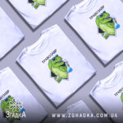 Футболка Травозавр білого кольору, зображена на молодому чоловікові, який позує з легким усмішкою. Принт на футболці зображає зеленого динозавра в окулярах, що тримає колбу з блакитною рідиною та пакет з травою. Фон світло-сірий, а текст 'ТРАВОЗАВР' розташований над принтом.