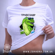 Футболка Травозавр білого кольору, розкладена на поверхні. Принт зображає зеленого динозавра в окулярах, що тримає колбу з блакитною рідиною та пакет з травою. На футболці є етикетка з логотипом, а фон світлий і однорідний.