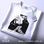 Футболка ти мій я твоя, біла футболка на плоскій поверхні, ракурс зверху, фон світлий, принт з чорними елементами, що зображує пару, текст на футболці: 'Ти мій... Я твоя... Ми разом - книга про життя...', з етикеткою зеленого кольору.