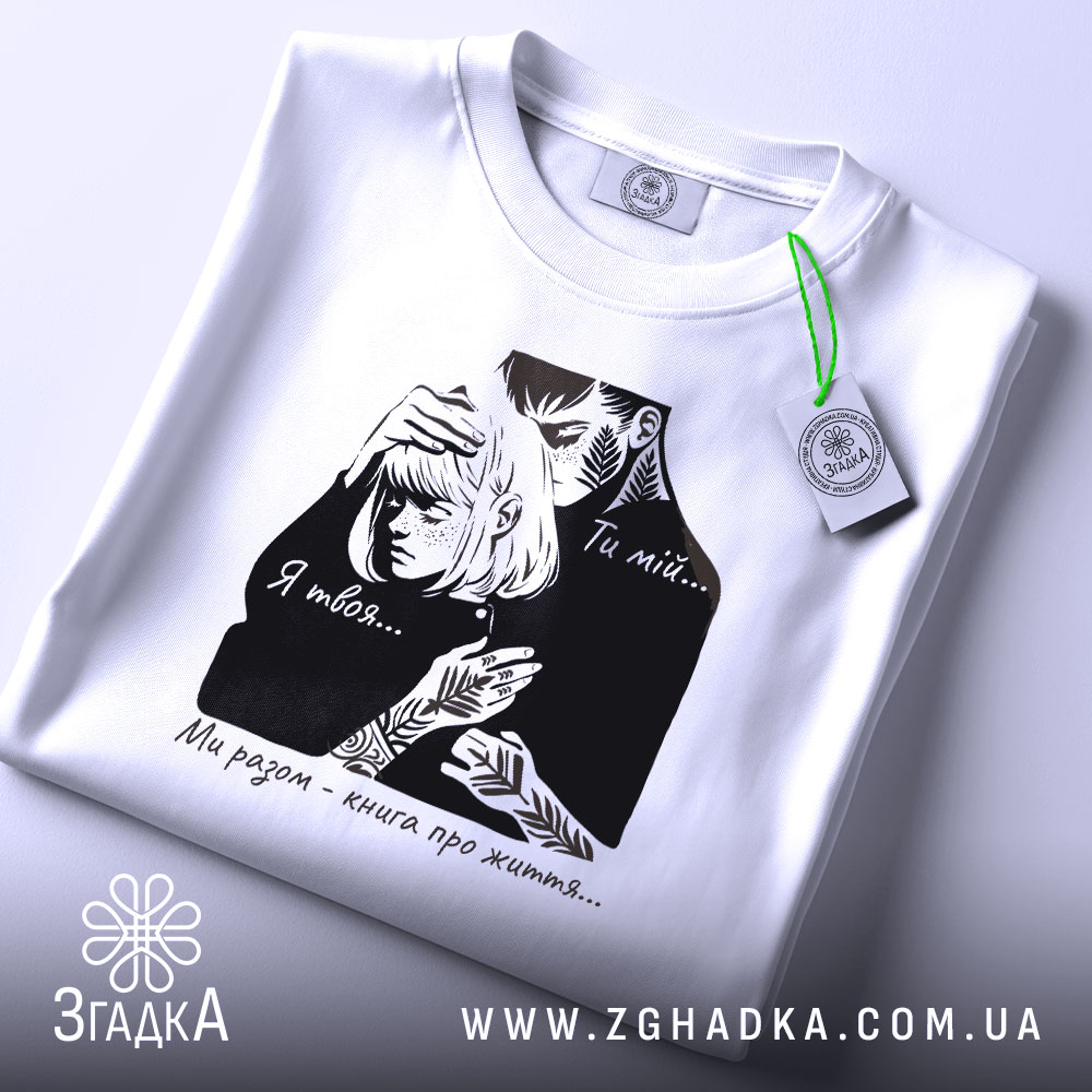 Футболка ти мій я твоя, біла футболка на плоскій поверхні, ракурс зверху, фон світлий, принт з чорними елементами, що зображує пару, текст на футболці: 'Ти мій... Я твоя... Ми разом - книга про життя...', з етикеткою зеленого кольору.