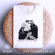 Футболка ти мій я твоя, біла футболка на плоскій поверхні, ракурс зверху, фон світлий, принт з чорними елементами, що зображує пару, текст на футболці: 'Ти мій... Я твоя... Ми разом - книга про життя...', на плетеній основі.