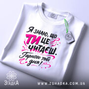 Футболка ти це читаєш, біла, з ракурсу зверху, на плоскій поверхні, фон світло-сірий, принт з текстом чорного та рожевого кольорів, з елементами графіки.