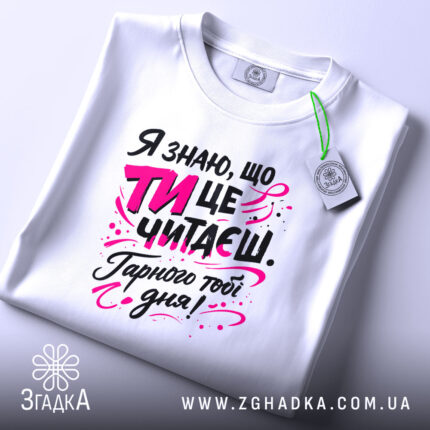 Футболка ти це читаєш, біла, з ракурсу зверху, на плоскій поверхні, фон світло-сірий, принт з текстом чорного та рожевого кольорів, з елементами графіки.