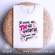 Футболка ти це читаєш, біла, з ракурсу зверху, на плоскій поверхні, фон світло-сірий, принт з текстом чорного та рожевого кольорів, розташована на плетеній основі.