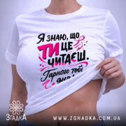 Футболка ти це читаєш, біла, з ракурсу зверху, на плоскій поверхні, фон світло-сірий, принт з текстом чорного та рожевого кольорів, з елементами графіки.