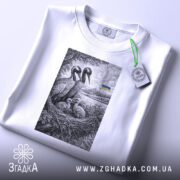 Футболка українські лелеки сімейний символ, біла, на плоскій поверхні, фон світло-сірий, принт з двома лелеками та пташенятами в гнізді, на задньому плані український прапор, етикетка з зеленим шнурком.