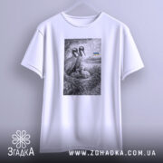 Футболка українські лелеки сімейний символ, біла, підвішена на вішалці, фон світло-сірий, принт з двома лелеками та пташенятами в гнізді, на задньому плані український прапор.