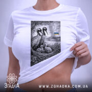 Футболка українські лелеки сімейний символ, біла, на плоскій поверхні, фон світло-сірий, принт з двома лелеками та пташенятами в гнізді, на задньому плані український прапор, етикетка з зеленим шнурком.