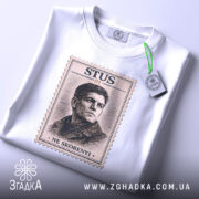 Футболка Василь Стус, біла, зображена на столі, ракурс зверху, фон світло-сірий, принт з портретом Василя Стуса в стилі поштової марки з написом 'STUS 01' та 'NE SKORENYI', на футболці є етикетка з зеленим шнурком.