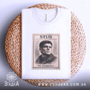 Футболка Василь Стус, біла, зображена на плетеній кошику, ракурс зверху, фон світло-сірий, принт з портретом Василя Стуса в стилі поштової марки з написом 'STUS 01' та 'NE SKORENYI'.