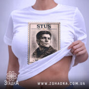 Футболка Василь Стус, біла, зображена на столі, ракурс зверху, фон світло-сірий, принт з портретом Василя Стуса в стилі поштової марки з написом 'STUS 01' та 'NE SKORENYI', на футболці є етикетка з зеленим шнурком.
