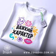 Футболка Важкий характер, біла, зображена зверху, на плоскій поверхні, принт яскравий, з елементами черепа, квітів та очей, текст "Важкий характер" великими чорними літерами, на комірці видно етикетку.