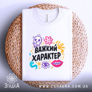 Футболка Важкий характер, біла, зображена зверху на плетеній кошику, фон світло-сірий, принт яскравий, з елементами черепа, квітів та очей, текст "Важкий характер" великими чорними літерами.