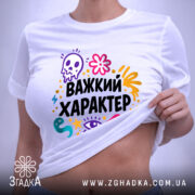 Футболка Важкий характер, біла, зображена зверху, на плоскій поверхні, принт яскравий, з елементами черепа, квітів та очей, текст "Важкий характер" великими чорними літерами, на комірці видно етикетку.