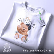 Футболка Ведмедик гангстер, біла, зображена на столі, ракурс зверху, фон світло-сірий, принт з ведмедиком у кепці та золотих ланцюгах, що тримає гроші, з етикеткою на комірці.