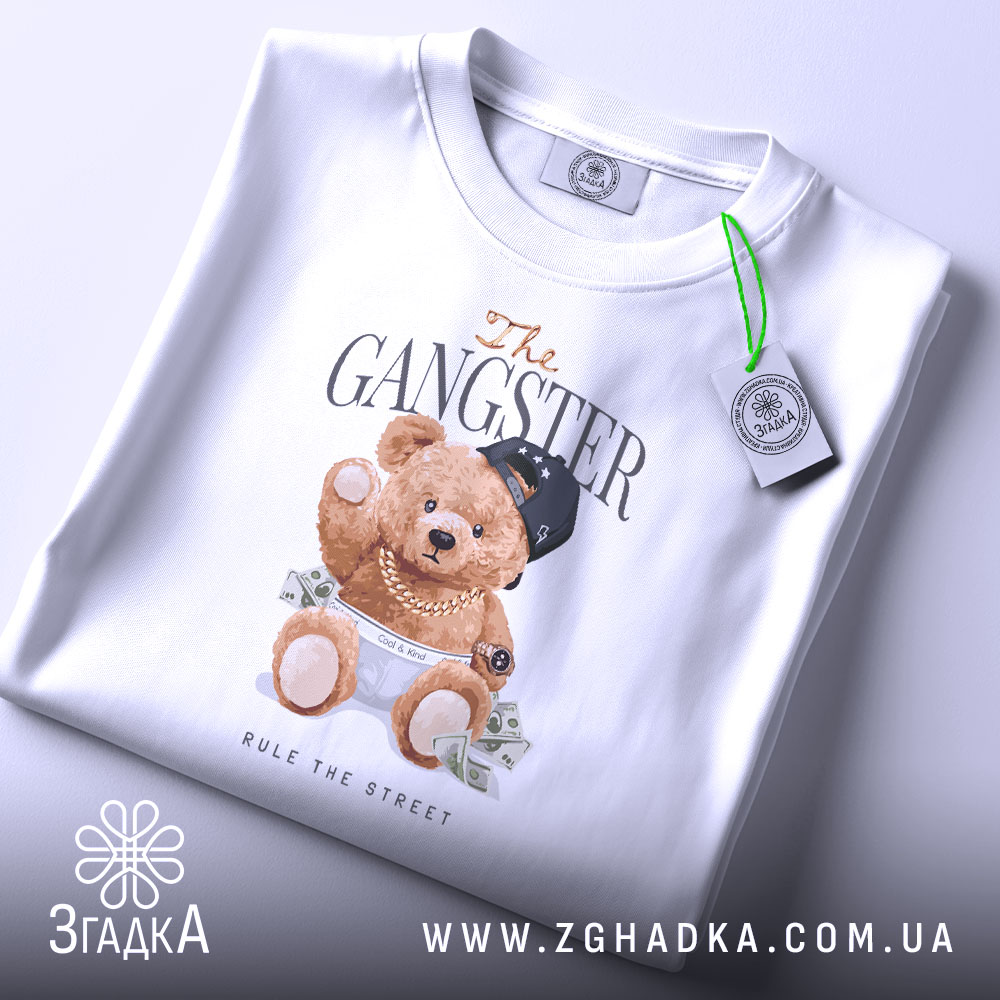 Футболка Ведмедик гангстер, біла, зображена на столі, ракурс зверху, фон світло-сірий, принт з ведмедиком у кепці та золотих ланцюгах, що тримає гроші, з етикеткою на комірці.