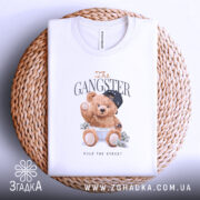 Футболка Ведмедик гангстер, біла, зображена на столі, ракурс зверху, фон плетений, принт з ведмедиком у кепці та золотих ланцюгах, що тримає гроші.