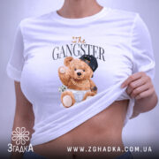 Футболка Ведмедик гангстер, біла, зображена на столі, ракурс зверху, фон світло-сірий, принт з ведмедиком у кепці та золотих ланцюгах, що тримає гроші, з етикеткою на комірці.