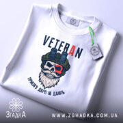 Футболка Veteran білого кольору, зображена на столі, ракурс зверху, фон світлий, принт з черепом у військовій касці та написом 'VETERAN', на футболці є етикетка з зеленим шнурком.