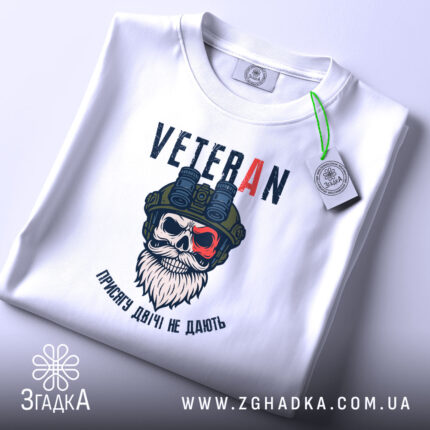 Футболка Veteran білого кольору, зображена на столі, ракурс зверху, фон світлий, принт з черепом у військовій касці та написом 'VETERAN', на футболці є етикетка з зеленим шнурком.