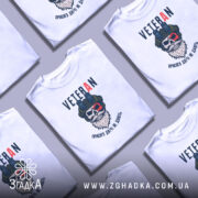 Футболка Veteran білого кольору, зображена на чоловікові, який позує в три чверті, фон світло-фіолетовий, принт з черепом у військовій касці та написом 'VETERAN' у верхній частині.