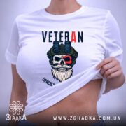 Футболка Veteran білого кольору, зображена на столі, ракурс зверху, фон світлий, принт з черепом у військовій касці та написом 'VETERAN', на футболці є етикетка з зеленим шнурком.