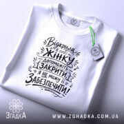 Футболка відкрила в собі жінку, біла футболка, розкладена на поверхні, фон світло-фіолетовий, принт чорного кольору з текстом на тему жіночої підтримки, з етикеткою зеленого кольору.