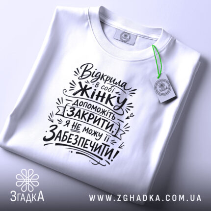 Футболка відкрила в собі жінку, біла футболка, розкладена на поверхні, фон світло-фіолетовий, принт чорного кольору з текстом на тему жіночої підтримки, з етикеткою зеленого кольору.