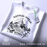 Футболка відповідь завжди чекає. Біла футболка, складена на поверхні, фон світло-фіолетовий. Принт з чорними елементами, що зображає гори, лижі та напис 'Чекає на вершині'. На футболці є етикетка з зеленим шнурком.