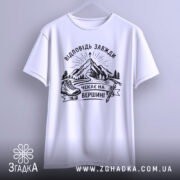 Футболка відповідь завжди чекає. Біла футболка, підвішена на вішалці, фон світло-сірий. Принт з чорними елементами, що зображає гори, лижі та напис 'Чекає на вершині'.