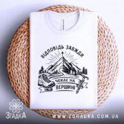 Футболка відповідь завжди чекає. Біла футболка, складена на плетеній кошику, фон світло-фіолетовий. Принт з чорними елементами, що зображає гори, лижі та напис 'Чекає на вершині'.