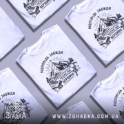 Футболка відповідь завжди чекає. Біла футболка на чоловікові, зображена в анфас, фон світло-фіолетовий. Принт з чорними елементами, що зображає гори, лижі та напис 'Чекає на вершині'.