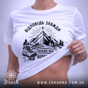 Футболка відповідь завжди чекає. Біла футболка, складена на поверхні, фон світло-фіолетовий. Принт з чорними елементами, що зображає гори, лижі та напис 'Чекає на вершині'. На футболці є етикетка з зеленим шнурком.