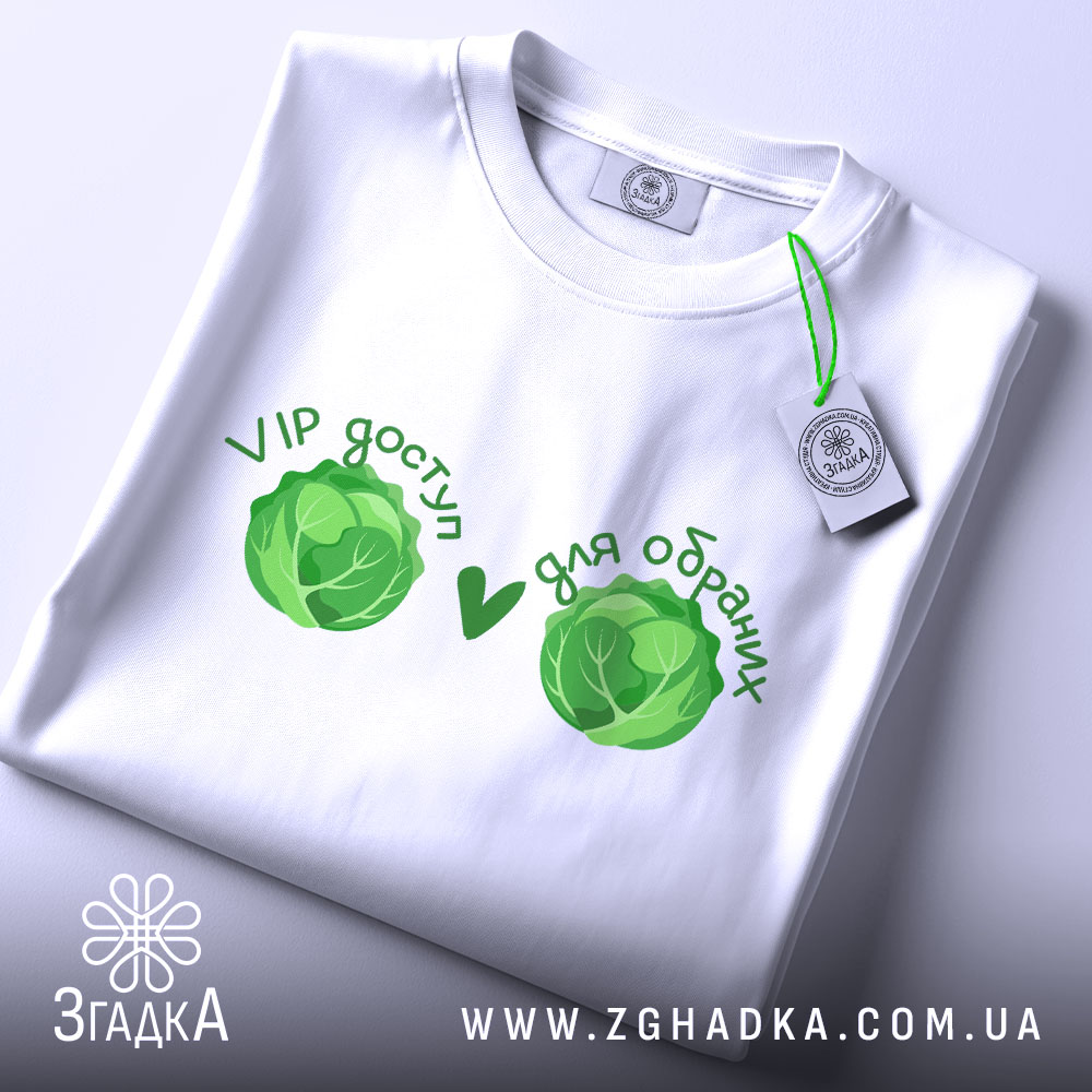 Футболка VIP доступ, біла, складена на столі, фон світлий, принт з двома зеленими капустами та текстом 'VIP доступ ♥ для обранців', видна етикетка з логотипом.