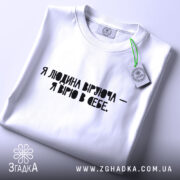 Футболка Віра в себе, біла, складена на поверхні, фон світло-сірий, принт чорного кольору з текстом "Я людина, що вірю - я вірю в себе", видна етикетка з логотипом.