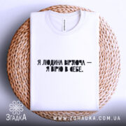 Футболка Віра в себе, біла, складена на плетеній основі, фон світло-сірий, принт чорного кольору з текстом "Я людина, що вірю - я вірю в себе".