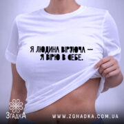 Футболка Віра в себе, біла, складена на поверхні, фон світло-сірий, принт чорного кольору з текстом "Я людина, що вірю - я вірю в себе", видна етикетка з логотипом.