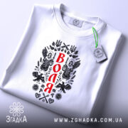 Футболка воля, складена на поверхні, білого кольору, з принтом на передній частині, зображення ангелів та герба України, фон світло-сірий, на футболці є етикетка з зеленим шнурком.
