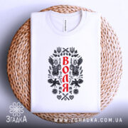 Футболка воля, складена на круглій кошику, білого кольору, з принтом на передній частині, зображення ангелів та герба України, фон світло-сірий, текст принта червоного кольору.