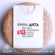 Футболка винна дієта, біла, складена на плетеній кошику, фон світло-сірий, принт чорний з червоним акцентом на слові 'БЕЗ', видима етикетка на комірці.