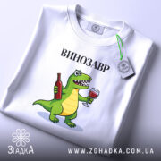 Футболка Винозавр, біла, на плоскій поверхні, з етикеткою, фон світло-сірий, принт з зеленим динозавром, що тримає пляшку вина та келих, текст 'ВИНОЗАВР' над принтом.