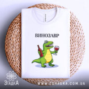 Футболка Винозавр, біла, складена на плоскій поверхні, фон світло-сірий, принт з зеленим динозавром, що тримає пляшку вина та келих, текст 'ВИНОЗАВР' над принтом.