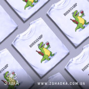 Футболка Винозавр, біла, на моделі, з легким макіяжем, фон світло-сірий, принт з веселим зеленим динозавром, який тримає в руках пляшку вина та келих, текст 'ВИНОЗАВР' над принтом.